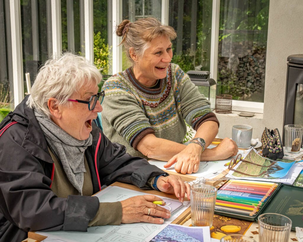 Kursister i Blomsterhaven To kursister i Blomsterhaven griner, mens de arbejder på deres haveplaner under et kursus i havedesign. De sidder ved et bord omgivet af planter og papir med skitser