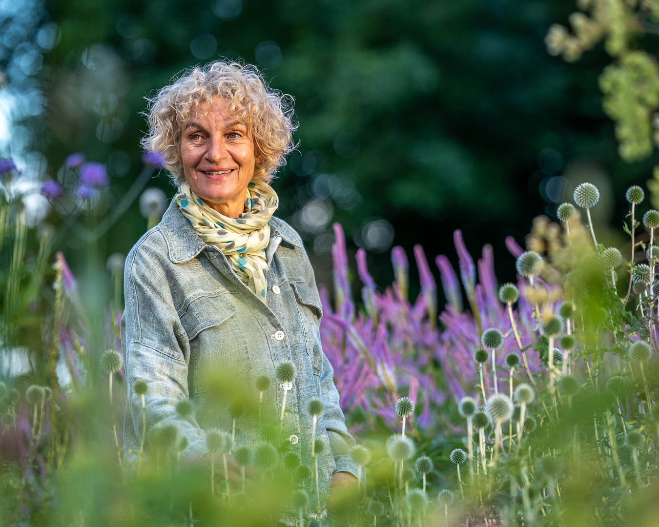 Portræt af Marianne Folling blandt lilla blomster og kugletidsler i hendes frodige have